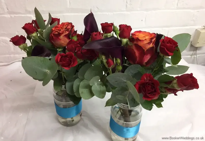 Table centrepiece jars of orange, raspberry & dark red coloured bouquet with roses, spray roses and calla lillies