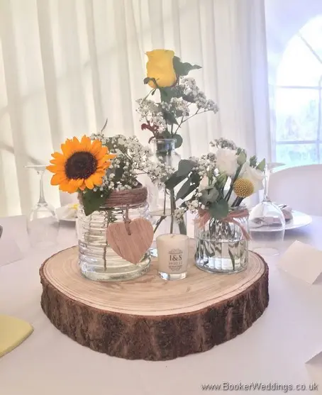 Table arrangements of 3 jars on rustic round log with yellow sunflowers, craspedia, roses, gypsy grass, lissanthus and grey eucalyptus