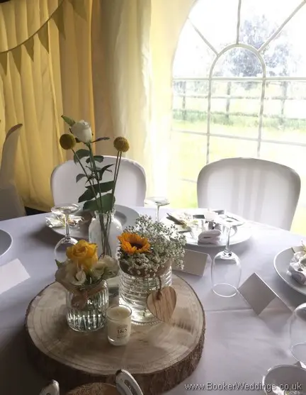 Table centrepieces of 3 jars on rustic round log with yellow sunflowers, craspedia, roses, gypsy grass, lissanthus and grey eucalyptus